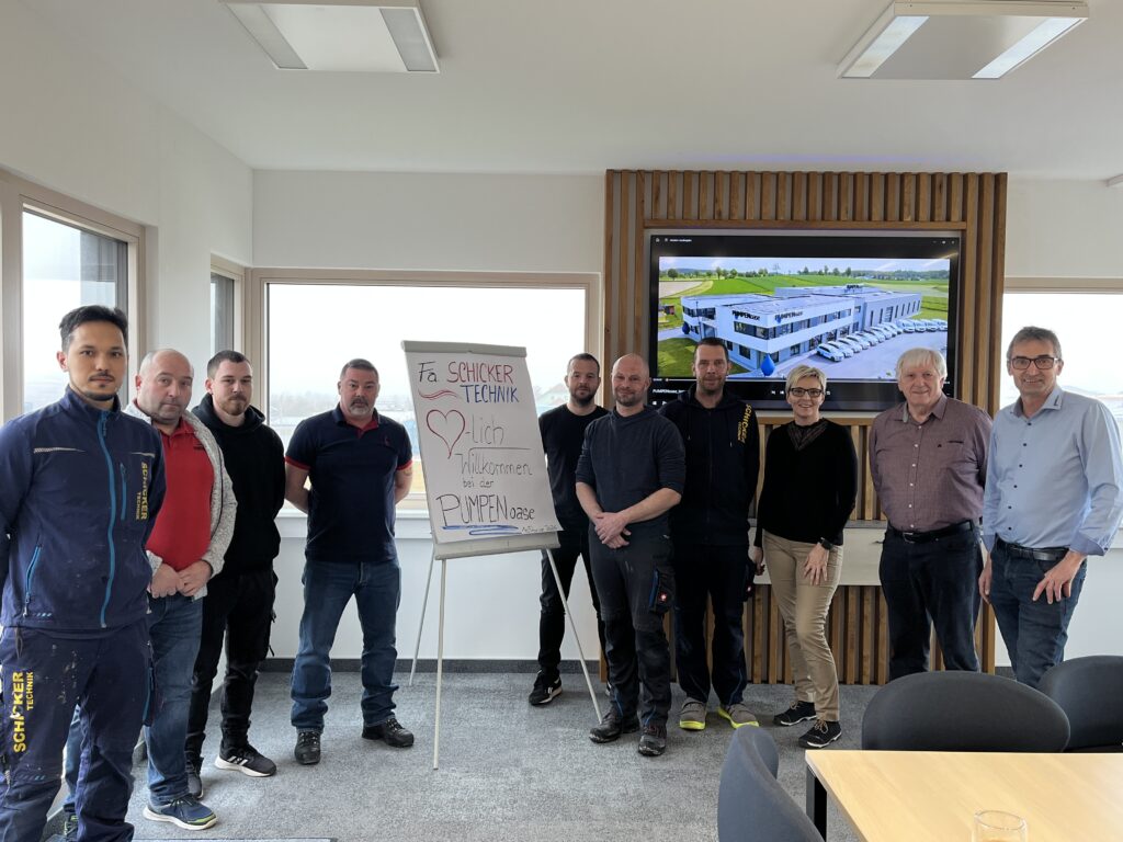 Team steht neben Begrüßungsplakat im Besprechungsraum.